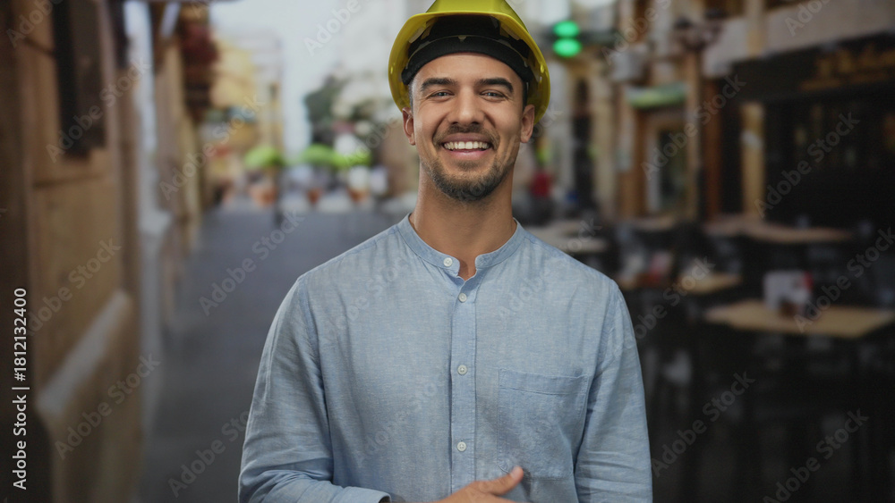 Fototapeta premium Young hispanic man with hardhat laughs on outdoor restaurant terrace, conveying joy and industry in a vibrant café street setting, blending work and leisure.