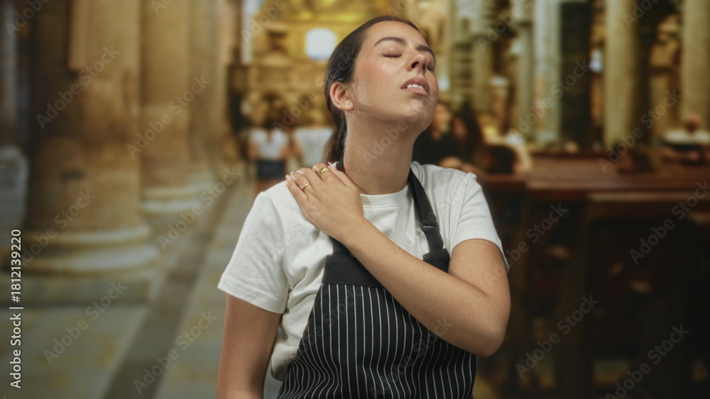 Fototapeta premium Young brunette woman in pinstripe apron holding hand to chest for heartattack inside church building while wincing in discomfort; faith struggle pain.