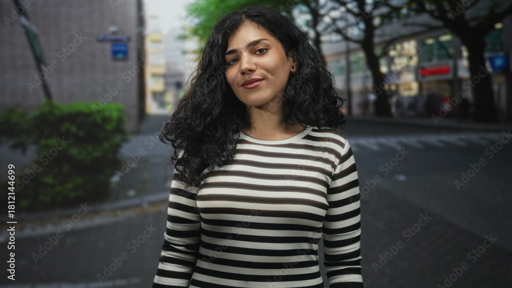 Fototapeta premium Young arab woman smiling with cheek piercings and striped top standing on a city street near trees and shops; confidence.