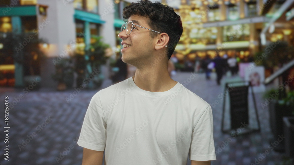 Fototapeta premium Man with curly hair and glasses turns his head to the side and smiles on a crowded city street, wearing a white t shirt; happiness.