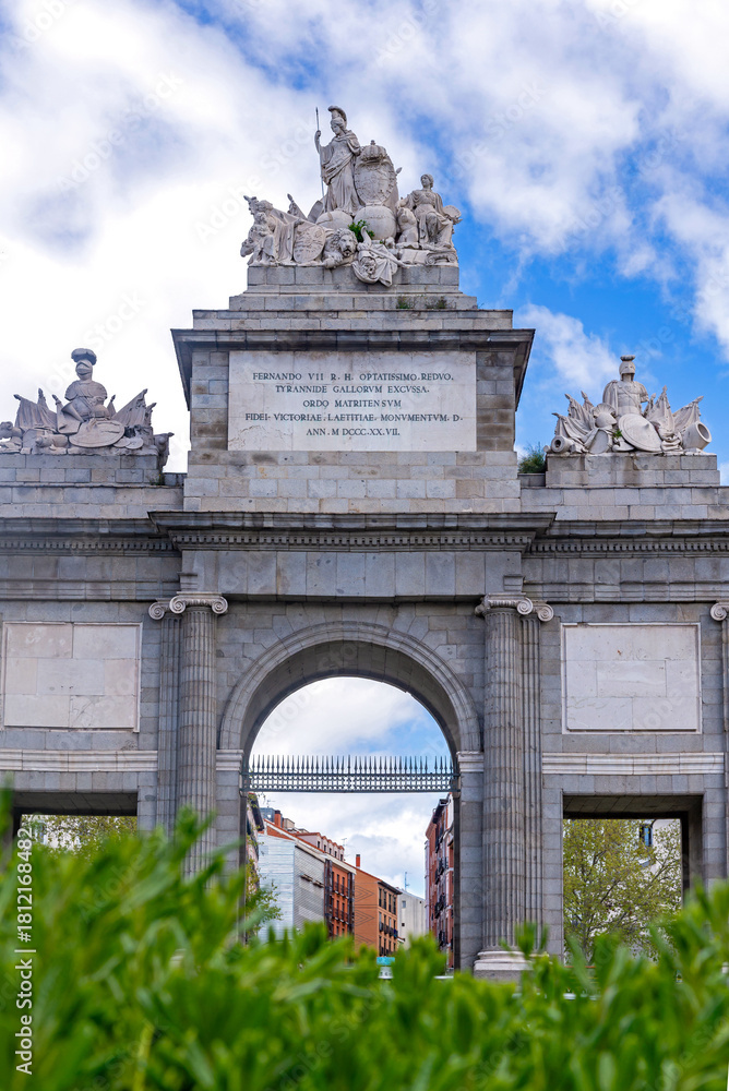 Obraz premium The Puerta de Toledo, a grand stone triumphal arch in Madrid, features three arches and sculptural groups at the center of a traffic circle under partly cloudy skies