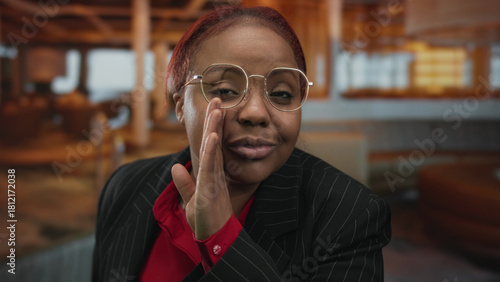 Obraz na plátně African american woman whispering with hand to mouth wearing round glasses in warm lit hotel building; secrecy
