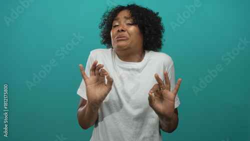 Quadro em tela Young african american woman wearing white t shirt cringing while holding ok sign in a teal studio; disgust