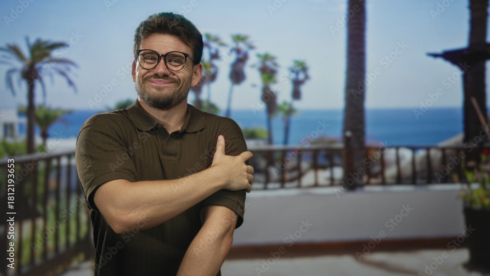 Obraz premium Man in brown polo shirt holds and rubs bare forearm on building balcony overlooking palm trees and ocean; discomfort seaside scene.