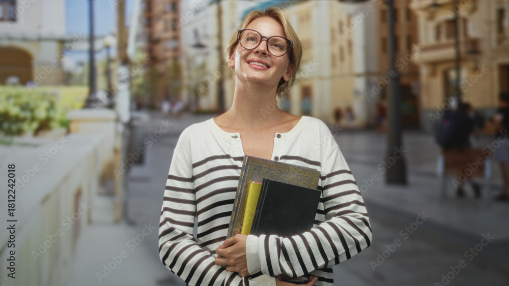 Fototapeta premium Blond woman with glasses holds books and a black notebook to her chest smiling with head tilt on a sunny city street lined with buildings and lampposts; joy study confidence.
