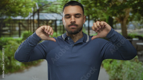 Man in blue polo shirt points thumbs to chest with closed eyes in green forest path setting; confidence.