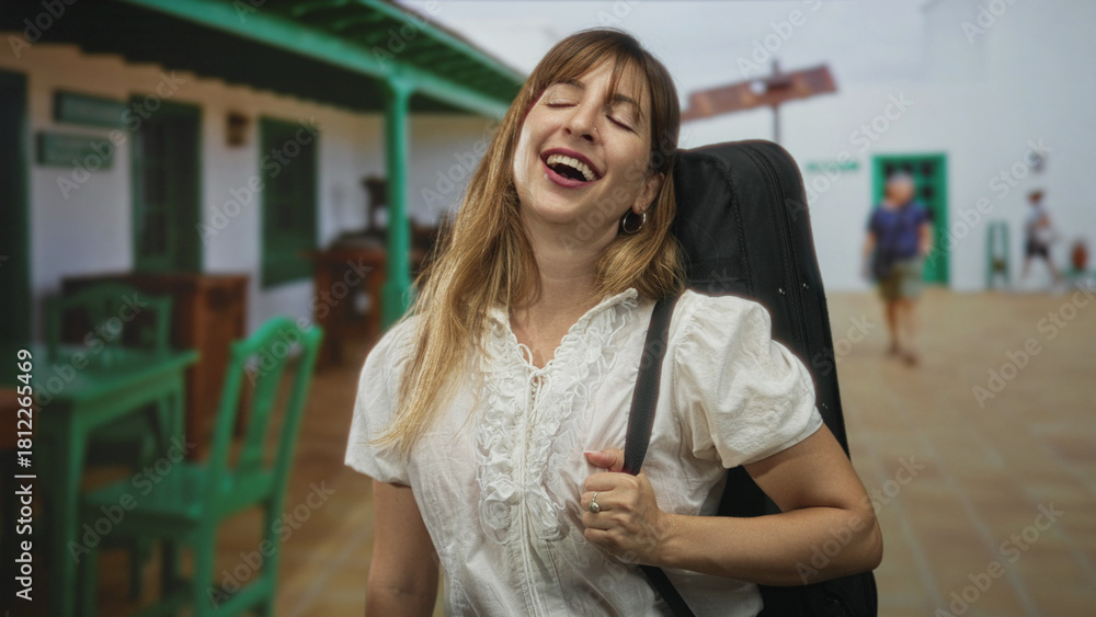 Fototapeta premium Woman carrying guitar case on shoulder holding strap and closing eyes while smiling in front of a white building porch with green chairs; joy travel.