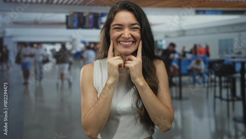 Free Download Young hispanic woman points finger to cheeks while puckering lips at busy airport terminal; playfulness. - GFXLoad