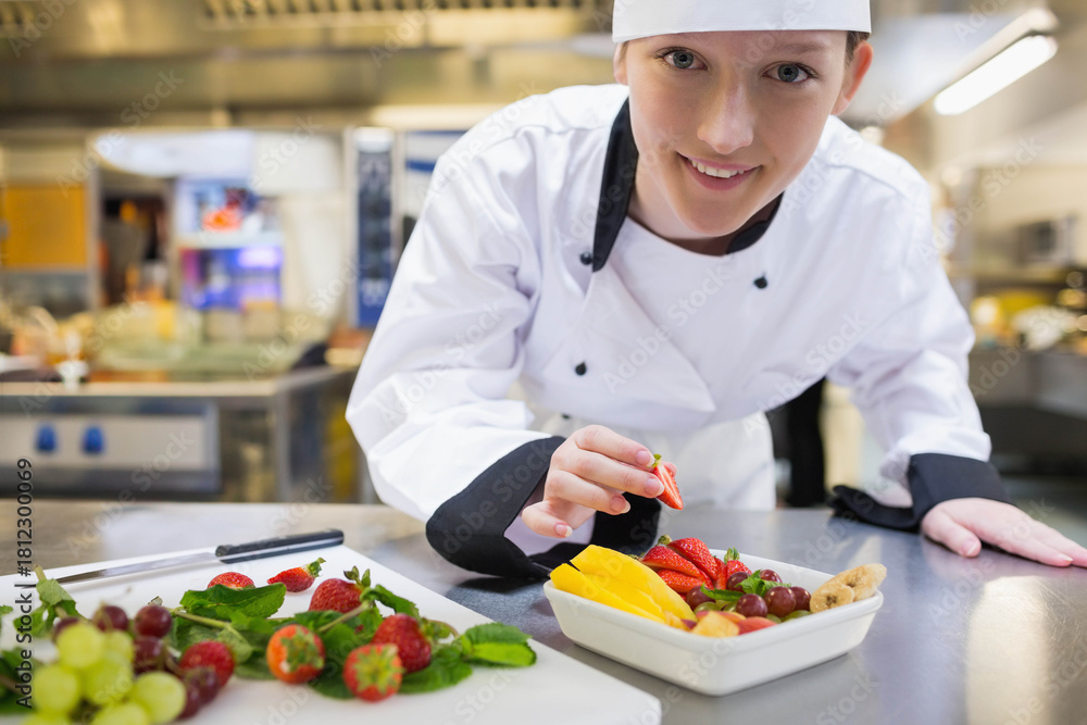 Naklejka premium Female chef wearing white chef coat leaning over counter in kitchen placing strawberry into dish