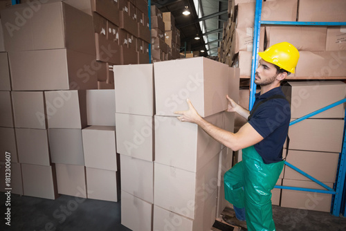 Male warehouse worker in yellow hard hat placing cardboard box onto stack near racking, copy space