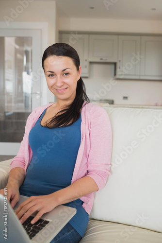 Free Download Adult woman sitting on couch in blue-top pink-cardigan typing on laptop at home, kitchen glass-door - GFXLoad