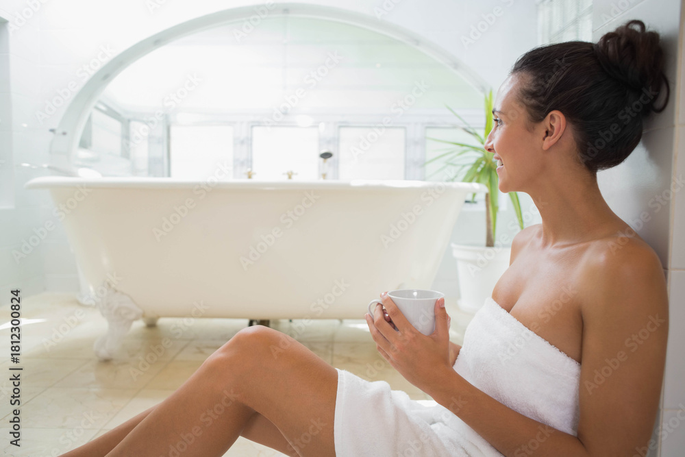 Obraz premium Adult woman sitting on bathroom floor wrapped in towel holding mug, facing clawfoot tub, copy space