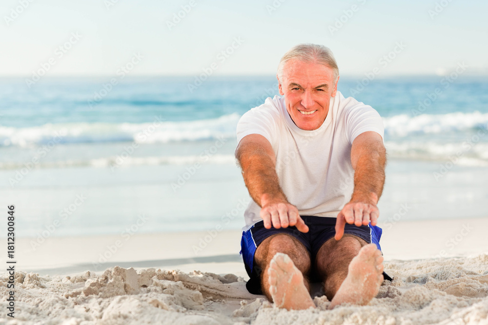 Fototapeta premium Senior man sitting and stretching on sandy beach near ocean waves wearing short-sleeve T-shirt