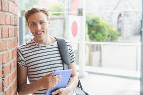 Man in twenties leaning against brick wall at entrance holding lavender tablet and gray bag smiling Free Download Man in twenties leaning against brick wall at entrance holding lavender tablet and gray bag smiling - GFXLoad