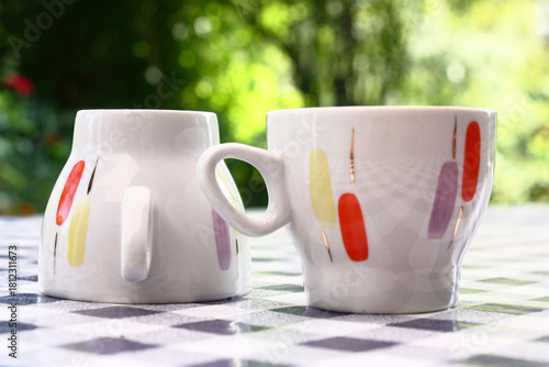 On a table with a checkered cloth there are two cups with original decorating. Garden plants serve as a green background.