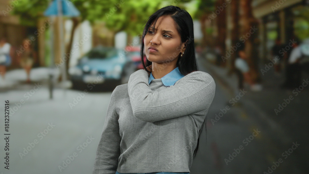 Fototapeta premium Young woman in gray sweater holds shoulder in discomfort on urban street with blurred background and trees lining the sidewalk.