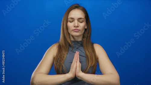 Standing woman with bare arms and hands pressed together in prayer in blue studio with calm gaze; serenity.