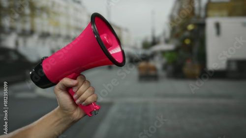 Man holding pink megaphone in city street signifies public announcement, showcasing hand raised with blurred background indicating urban action and spontaneous speech.