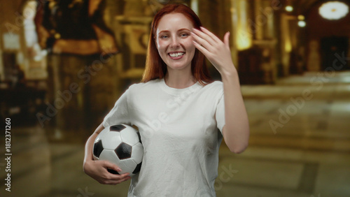 Woman standing in a church holding a soccer ball, smiling with hand covering eyes, suggesting joy and diversion within a sacred setting.