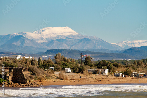 Fototapeta Naklejka Na Ścianę i Meble -  Storm waves crash on a sandy beach near ancient fortress ruins under sunny skies. Snow-capped mountains rise in the distance behind deserted shrubland. A dramatic Mediterranean scene in Side, Тurkey,