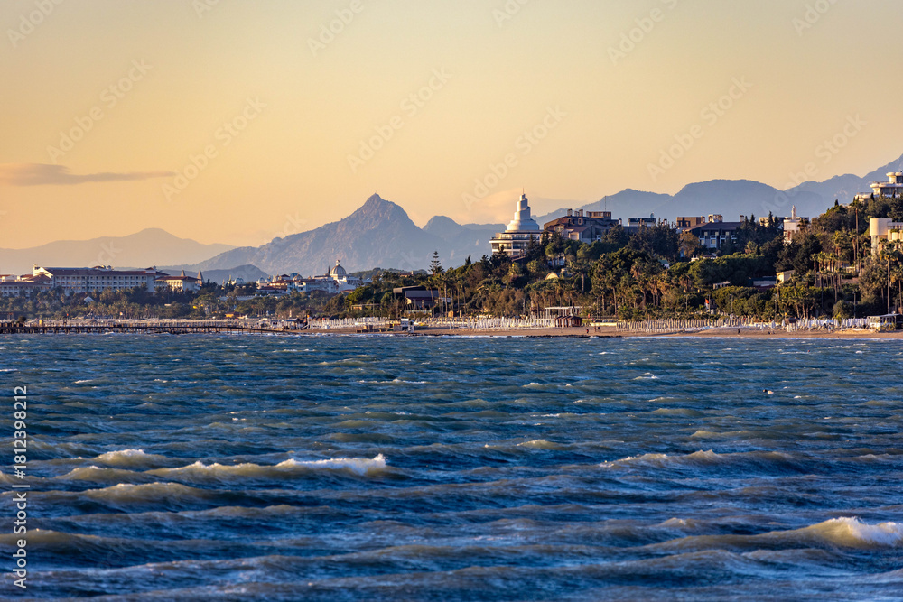 Naklejka premium Wavy blue sea with whitecaps under evening sun, viewed from the water towards a resort coastline with beach umbrellas, hotels, and misty mountains. Side, Antalya, Turkey, Mediterranean.