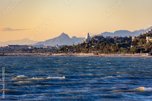 Fototapeta Naklejka Na Ścianę i Meble -  Wavy blue sea with whitecaps under evening sun, viewed from the water towards a resort coastline with beach umbrellas, hotels, and misty mountains. Side, Antalya, Turkey, Mediterranean.

