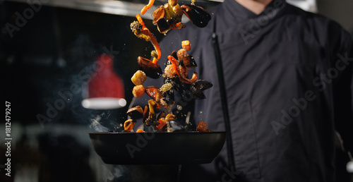 Tablou pe pânză Chef tossing sauteed vegetables in pan over stove in kitchen, dark background