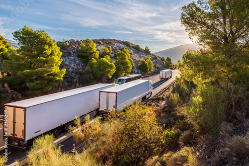 Several trucks driving along a two-way mountain road bordered by dense forests.