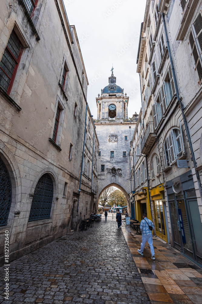 Obraz premium A street view in La Rochelle looking towards the iconic Grosse Horloge clock tower gate. Wet cobblestones and historic stone buildings frame the medieval archway under an overcast sky.