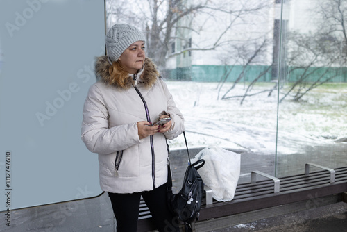 A woman sits at a bus stop in winter. While waiting for the bus, she has a bag and a dog in her hands.