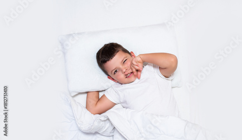 A young boy is lying in bed on white pillows and white bedding. He is smiling brightly as if he has just woken up. The boy looks happy, relaxed, and comfortable