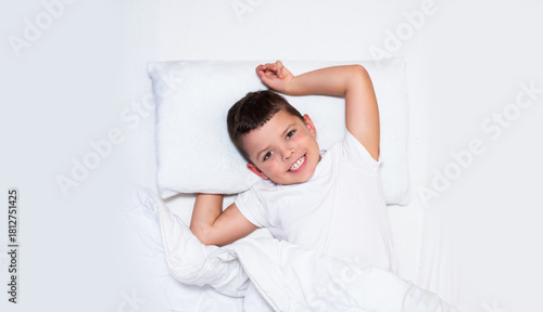 A young boy is lying in bed on white pillows and white bedding. He is smiling brightly and stretching his arms above his head, as if he has just woken up. The boy looks happy, relaxed, and comfortable
