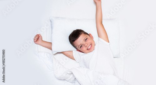 A young boy is lying in bed on white pillows and white bedding. He is smiling brightly and stretching his arms above his head, as if he has just woken up. The boy looks happy, relaxed, and comfortable