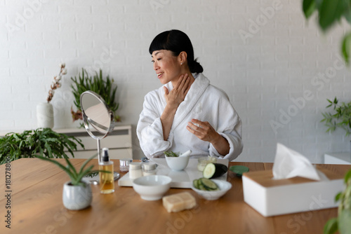 Asian woman in a bathrobe touching her neck while preparing skincare products during a calm home beauty routine at a wooden table.