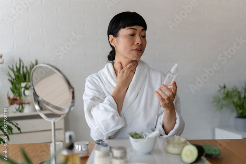 Asian woman in a bathrobe examining a skincare serum bottle while touching her neck during a calm home beauty routine at a table