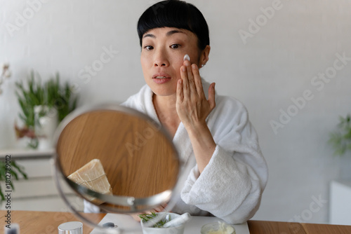 Quadro em tela Asian woman in a white bathrobe applying face cream to her cheek, captured in a