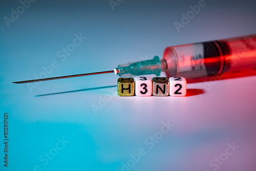 Syringe with red liquid and lettered blocks spelling H3N2, symbolizing vaccination and health awareness, set against a gradient background with soft lighting effects