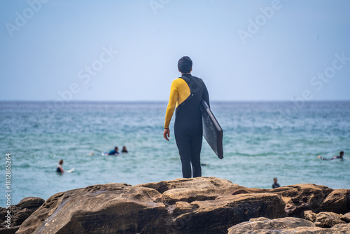 Surfer in wetsuit holding board looks out at the ocean with other surfers in the water