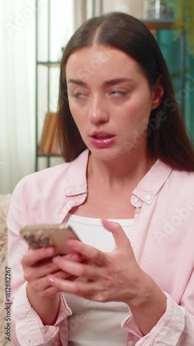 Young woman sitting on home sofa angrily types on smartphone chatting and waves hand in frustration at negative messages. Caucasian girl shows displeased look and posture reacting to words online.