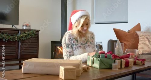 Woman wearing Santa hat wrapping gifts tying red ribbon at living room table by wood-burning stove