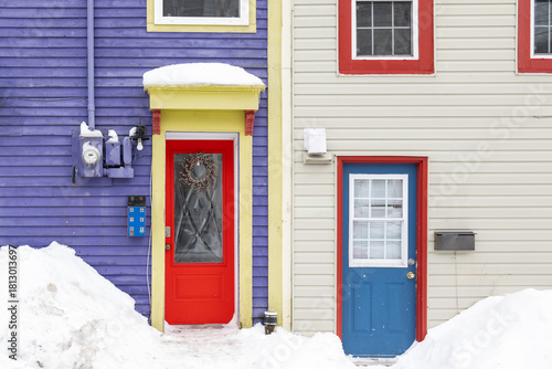 Two colorful doors on neighboring houses. There's a purple wooden house with a red door and glass windows. There's bright yellow trim around the door. The other house is white with a blue door.