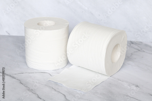 Two rolls of white toilet paper, with one partially unrolled, standing on a clean marble surface, symbolizing hygiene and household essentials.