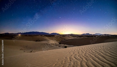 Fototapeta Naklejka Na Ścianę i Meble -  A serene nightscape shows rolling desert dunes under a starry sky. The horizon glows faintly, silhouetting mountain range