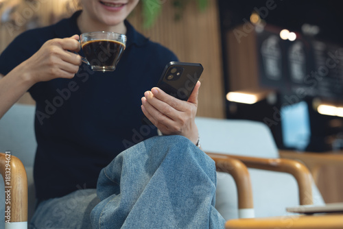 Happy young asian woman using smartphone and drinking coffee, social network at coffee shop. Smiling freelancer using mobile phone video call at cafe, modern people lifestyle