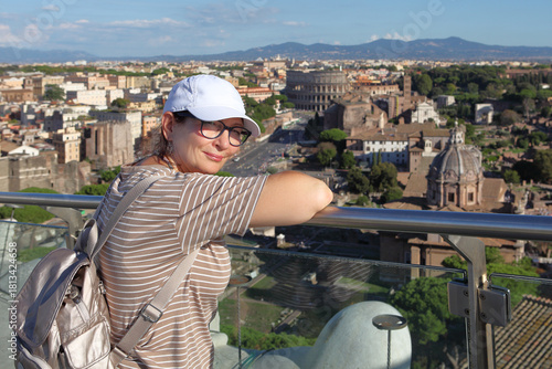 Tourist woman enjoying a panoramic view of the ancient Rome skyline from a high terrace, featuring the iconic Colosseum and Roman Forum ruins under a bright blue sky.