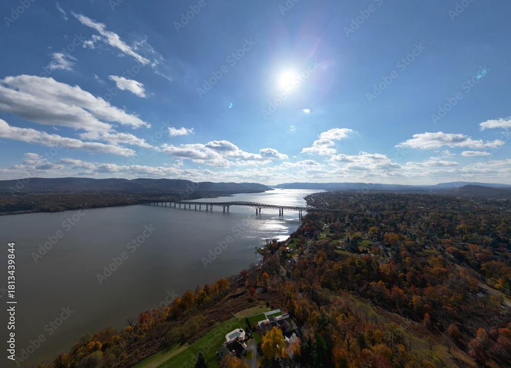 Obraz premium Aerial view of Hudson River with strong sunlight reflecting off the water near Newburgh, NY, showcasing surrounding autumn foliage and deep blue skies. Travel photography.