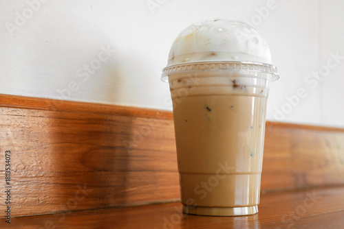 Iced coffee drink in clear plastic cup with dome lid and milk foam on wooden table in cafe.