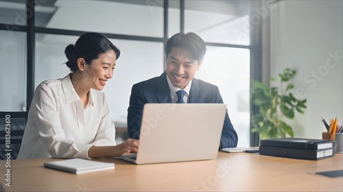 Two Asian business colleagues working together on a laptop in a modern office, collaborating on a project with smiles and positive energy.