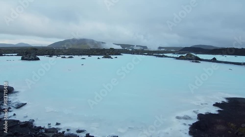 An aerial or wide shot of the famous Blue Lagoon geothermal spa in Iceland, showing the milky blue water surrounded by dark volcanic lava fields. In the background, the Svartsengi Geothermal Power Pla
