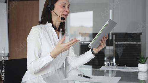 Businesswoman in a white shirt and headset leading a video call from a modern office, presenting with a tablet and gesturing as she discusses strategy and trains remote colleagues